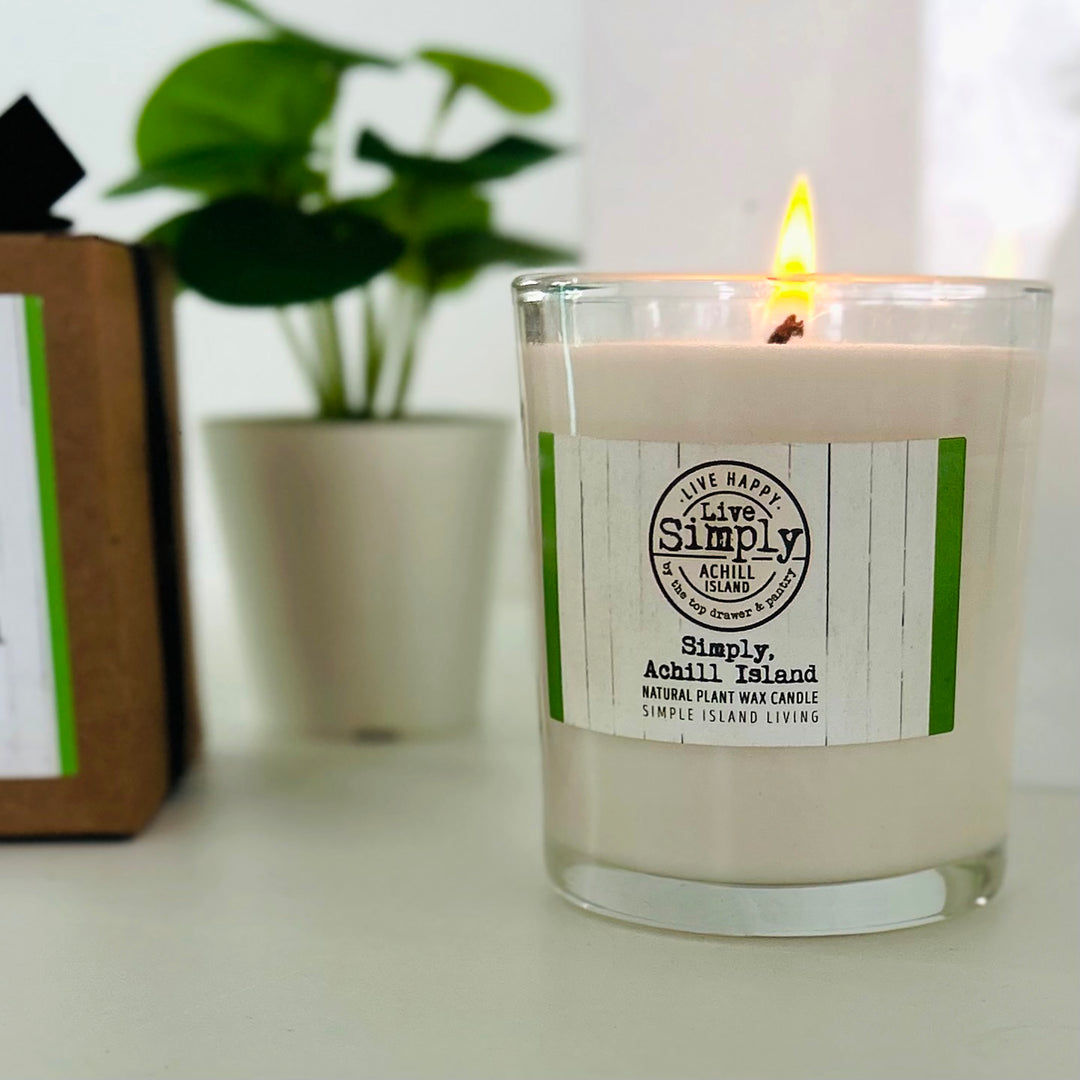 Lit Simply Achill Island candle beside its kraft gift box, with a leafy plant in the background