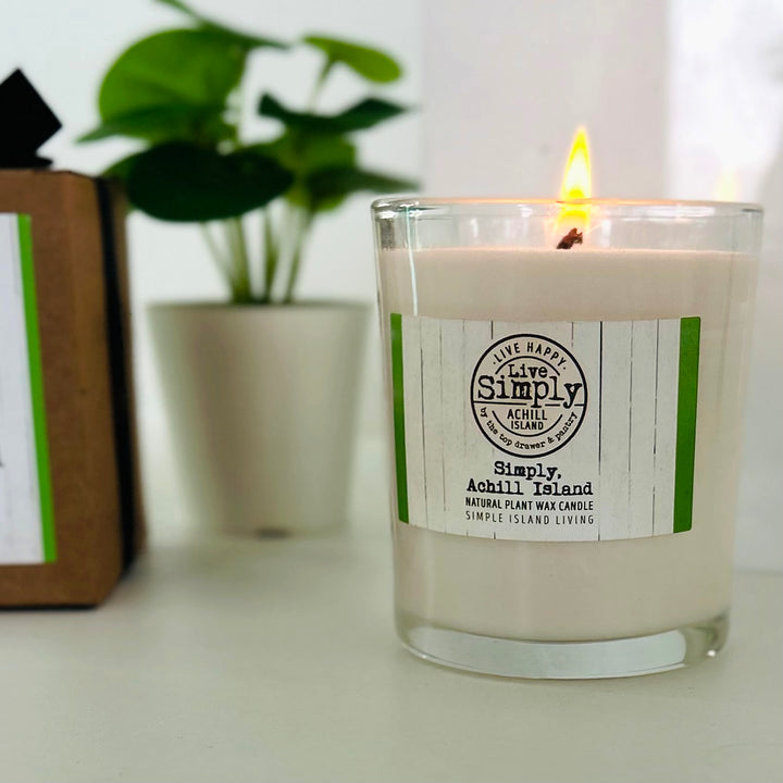 Lit Simply Achill Island candle beside its kraft gift box, with a leafy plant in the background