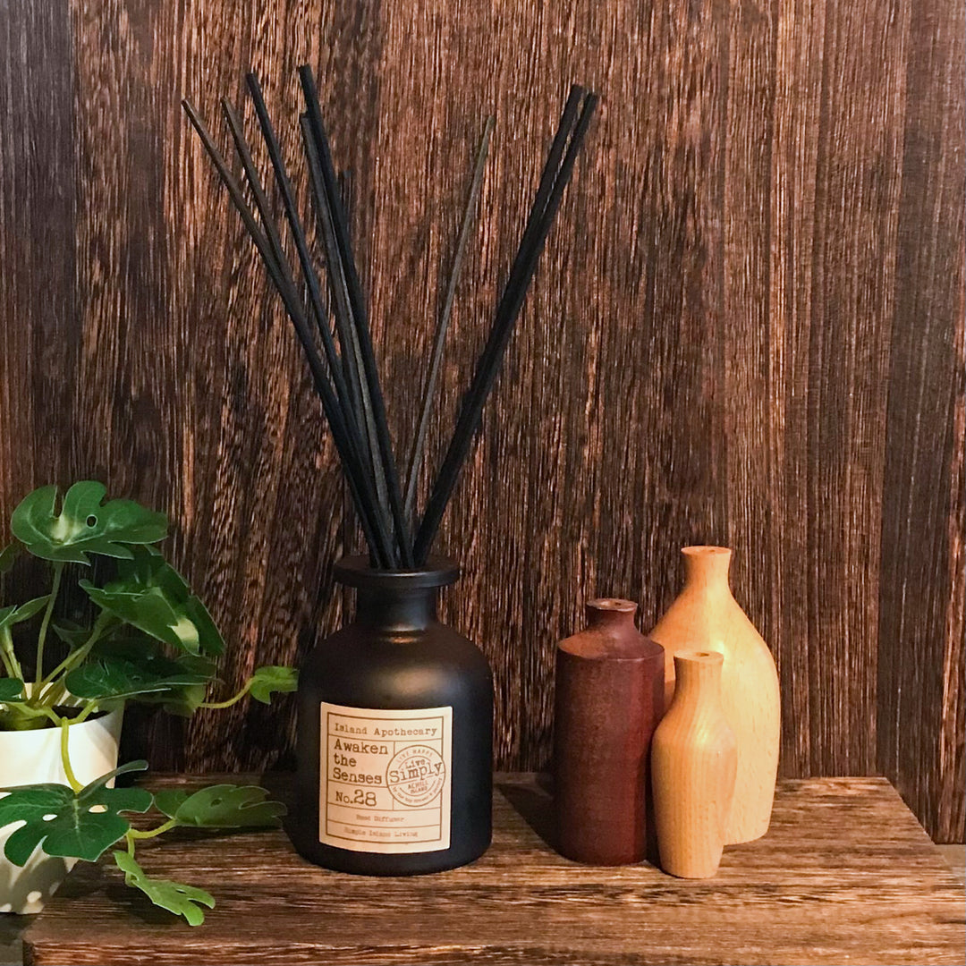 Black vase with black sticks on a wooden surface with other vases and a plant in the background.