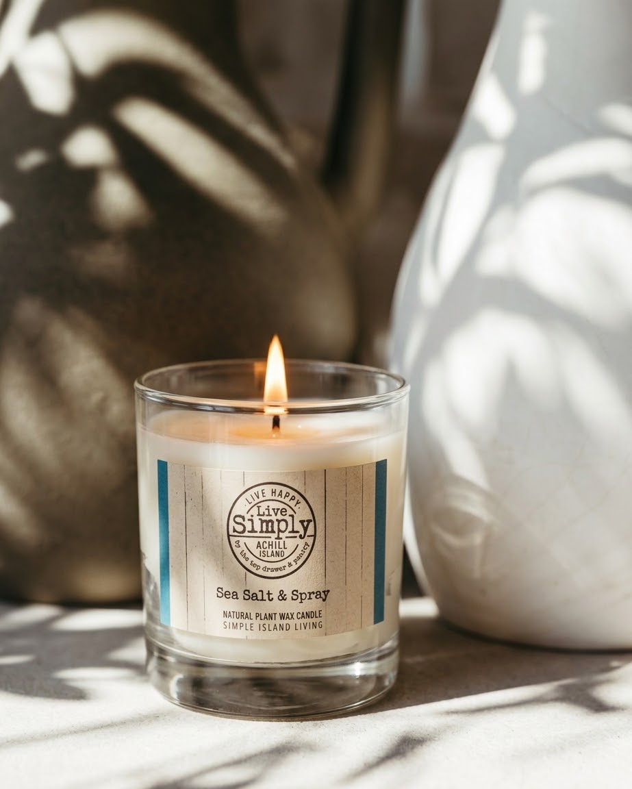 A lit candle in a clear glass jar with a cream-coloured label reading 'Sea Salt & Spray.' The candle sits on a light surface with strong dappled shadows, and a white ceramic vase is blurred in the background.