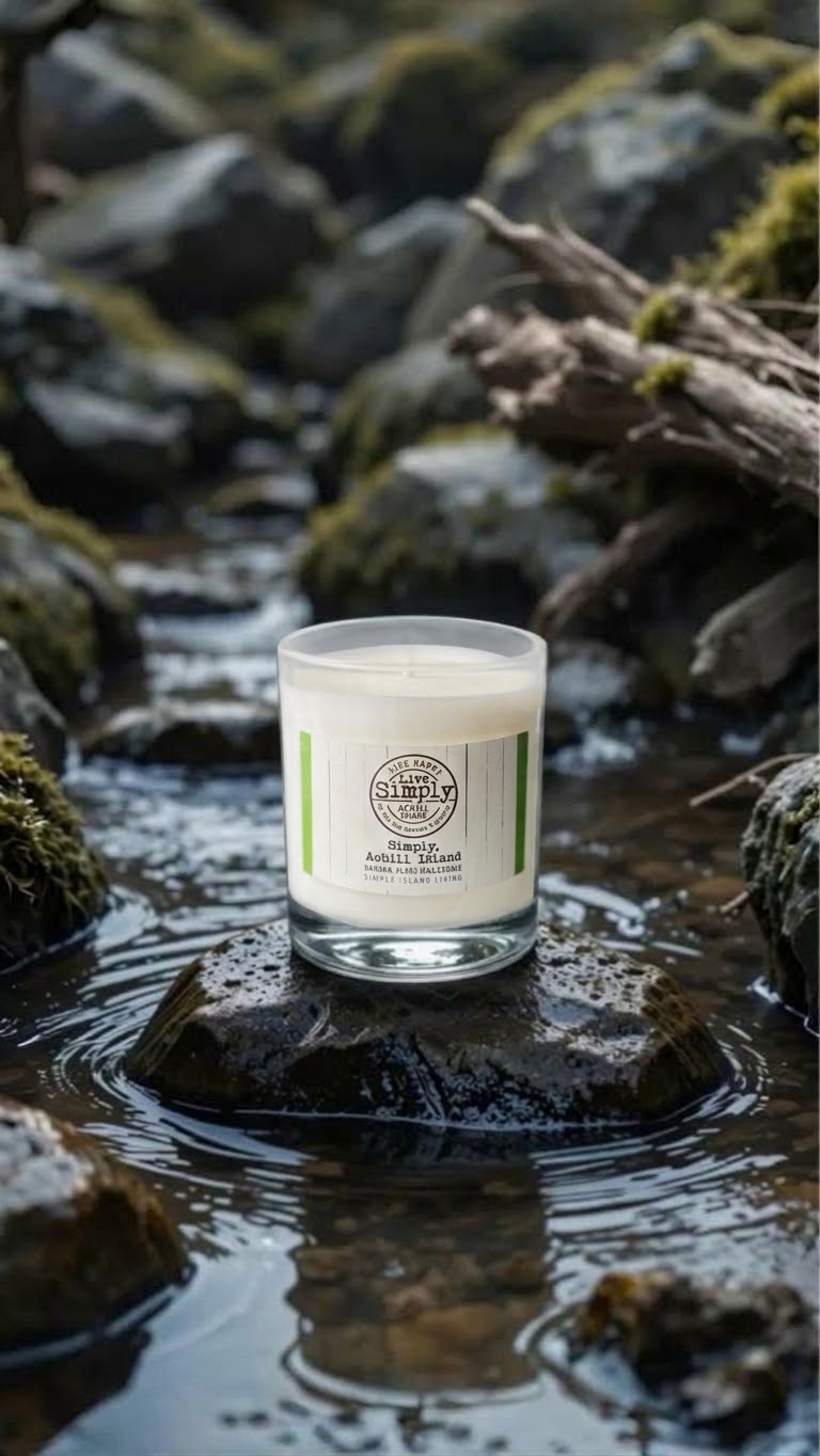 A glass jar candle with white wax sits on a wet rock in a shallow stream, surrounded by mossy rocks and water reflections. The label reads simply Achill Island 